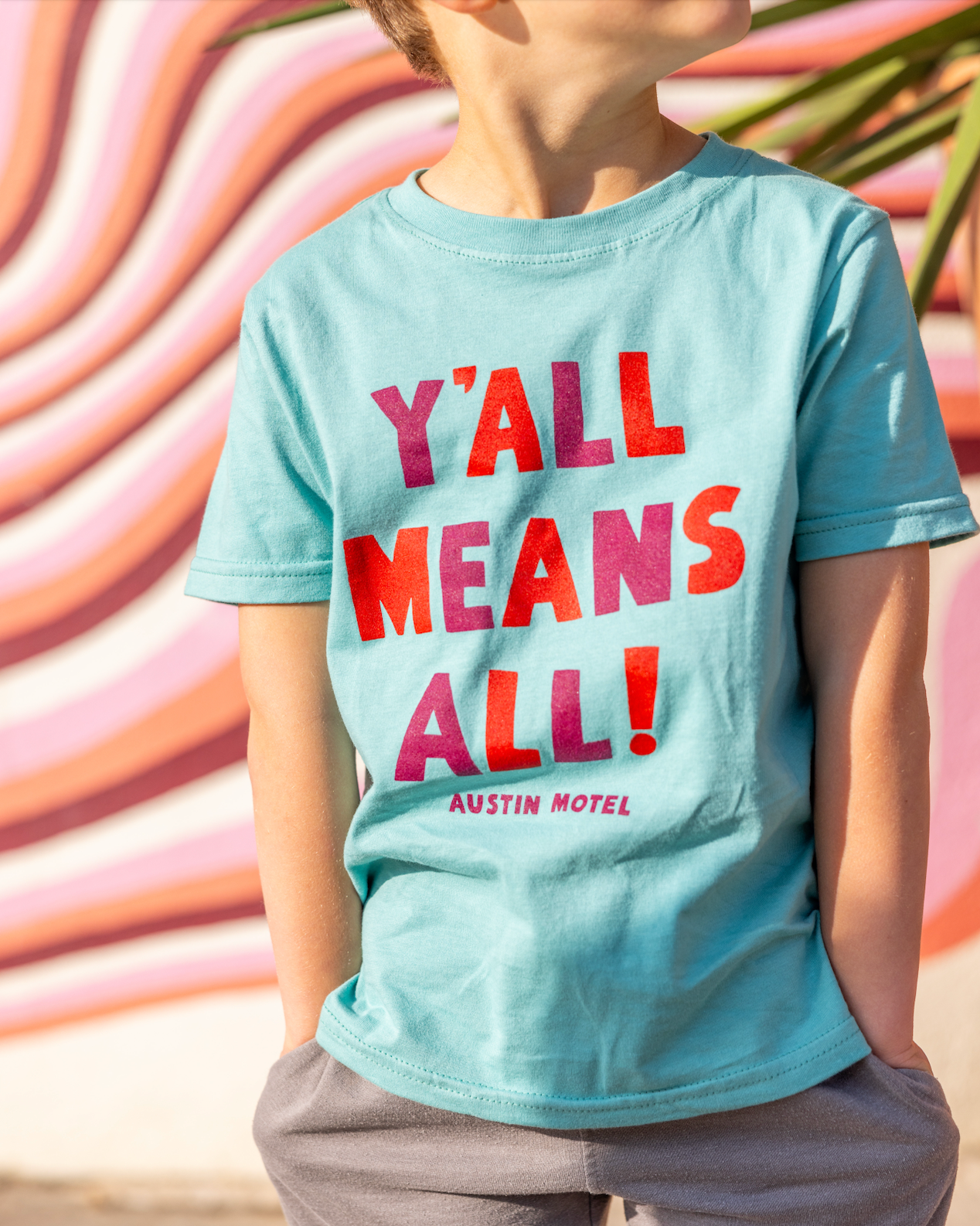 Child wearing a light blue t-shirt with colorful text that reads 'YALL MEANS ALL' in front of a wavy, multicolored background.