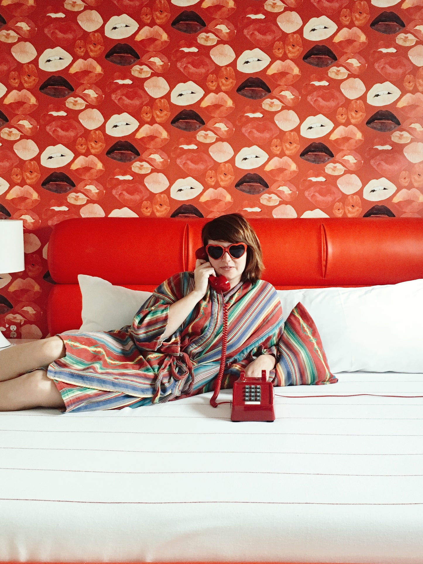 Woman lounging on Austin Motel bed wearing cotton striped serape robe and heart shaped sunglasses holding a cord phone to her ear