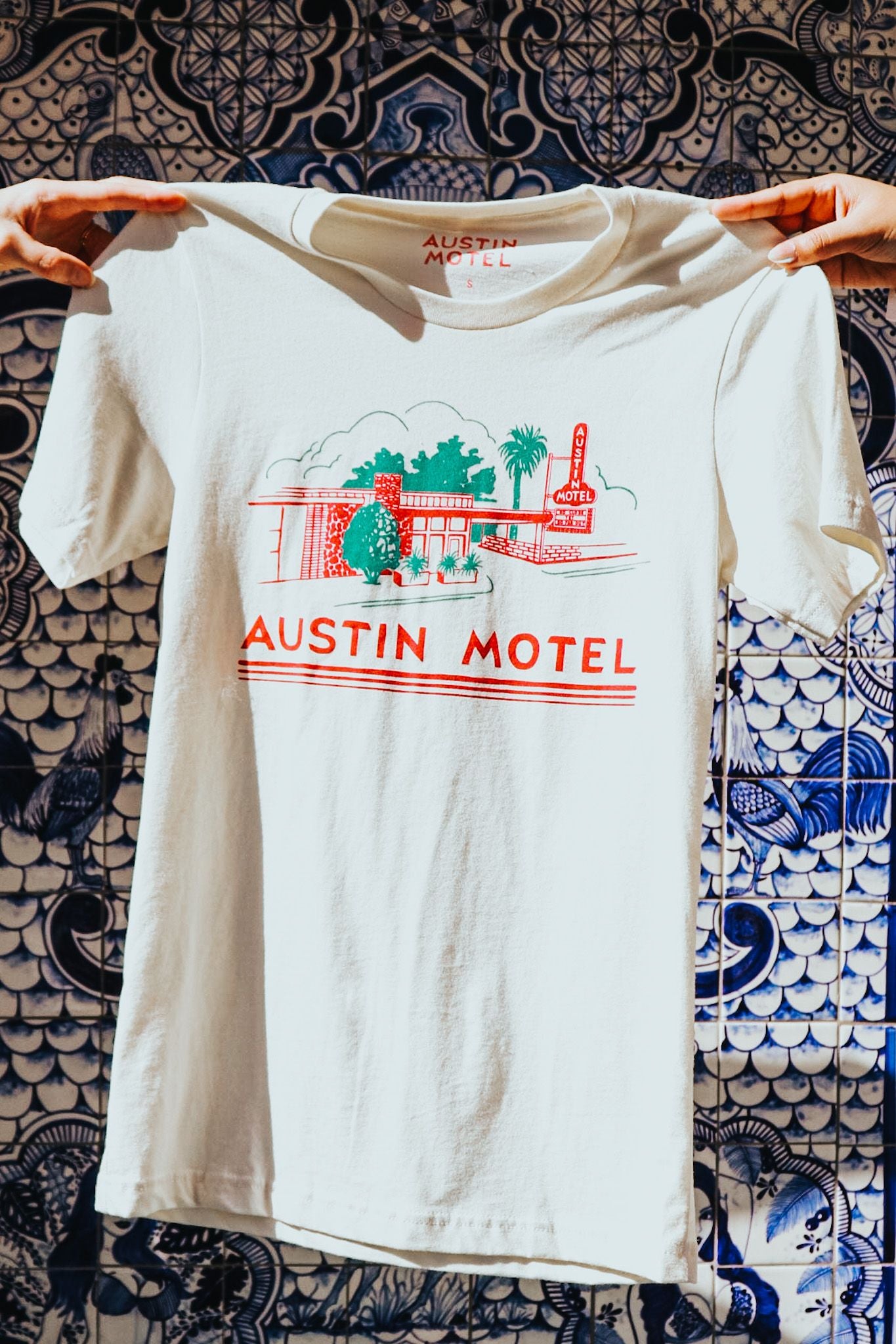 A cream t-shirt with a colorful illustration of the Austin Motel building and the text 'AUSTIN MOTEL' printed across the chest being held by two hands on either side in front of a decorative hand painted tile wall.