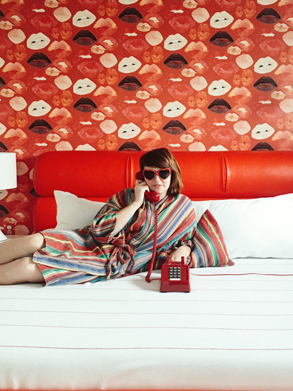 Woman lounging on Austin Motel bed wearing cotton striped serape robe and heart shaped sunglasses holding a cord phone to her ear