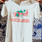A cream t-shirt with a colorful illustration of the Austin Motel building and the text 'AUSTIN MOTEL' printed across the chest being held by two hands on either side in front of a decorative hand painted tile wall.